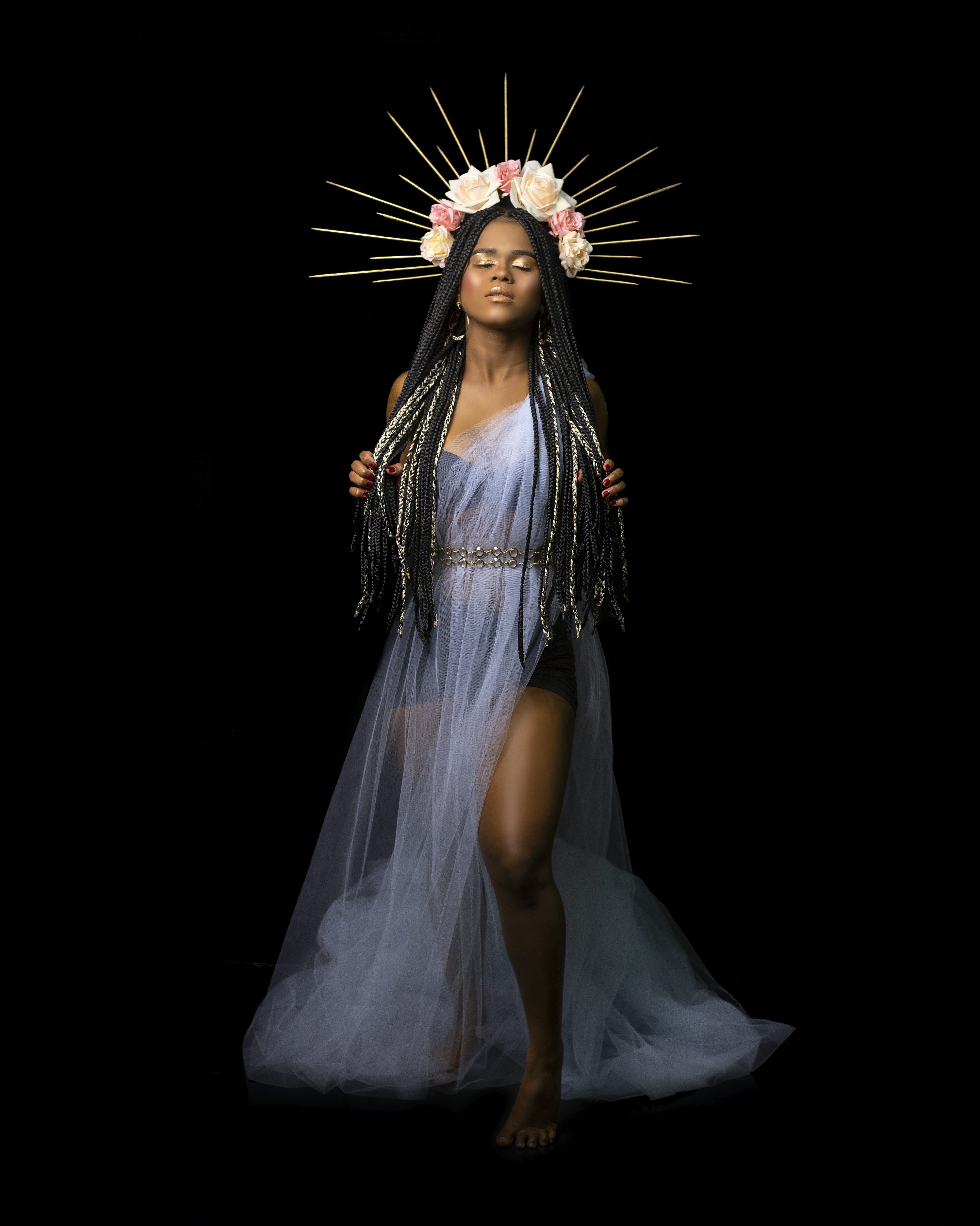 a black woman with long box braids is captured in a studio shot against a solid black background. she is adorned with a beautiful flower crown composed of pastel pink and white roses, accented with a golden sunburstlike halo.  she wears a flowing, sheer, lightblue tulle dress that accentuates her graceful pose. her eyes are closed, and she exudes a serene and spiritual aura.  the overall image conveys a sense of ethereal beauty, power, and selfassuredness.