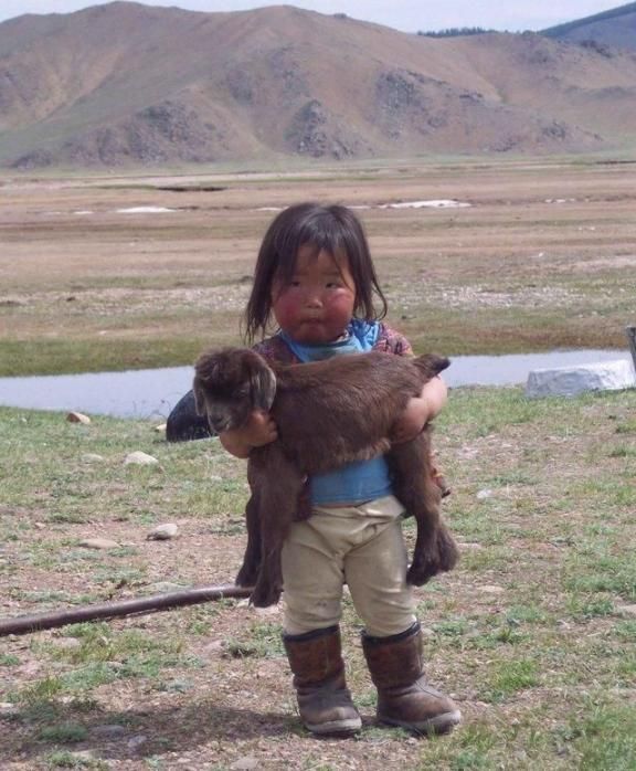 a young child with rosy cheeks is holding a baby goat in a rural outdoor setting. the child is wearing lightcolored pants and brown boots. behind them is a landscape with a river and mountains.