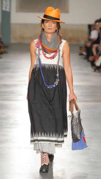 a fashion model strolls down a runway wearing a black dress and a scarf, she is accessorizing her outfit with a hat and a bag and black boots. the outfit has a  boho chic vibe with colorful details.  her pose is elegant and stylish and the outfit perfectly embodies the latest fashion trends.