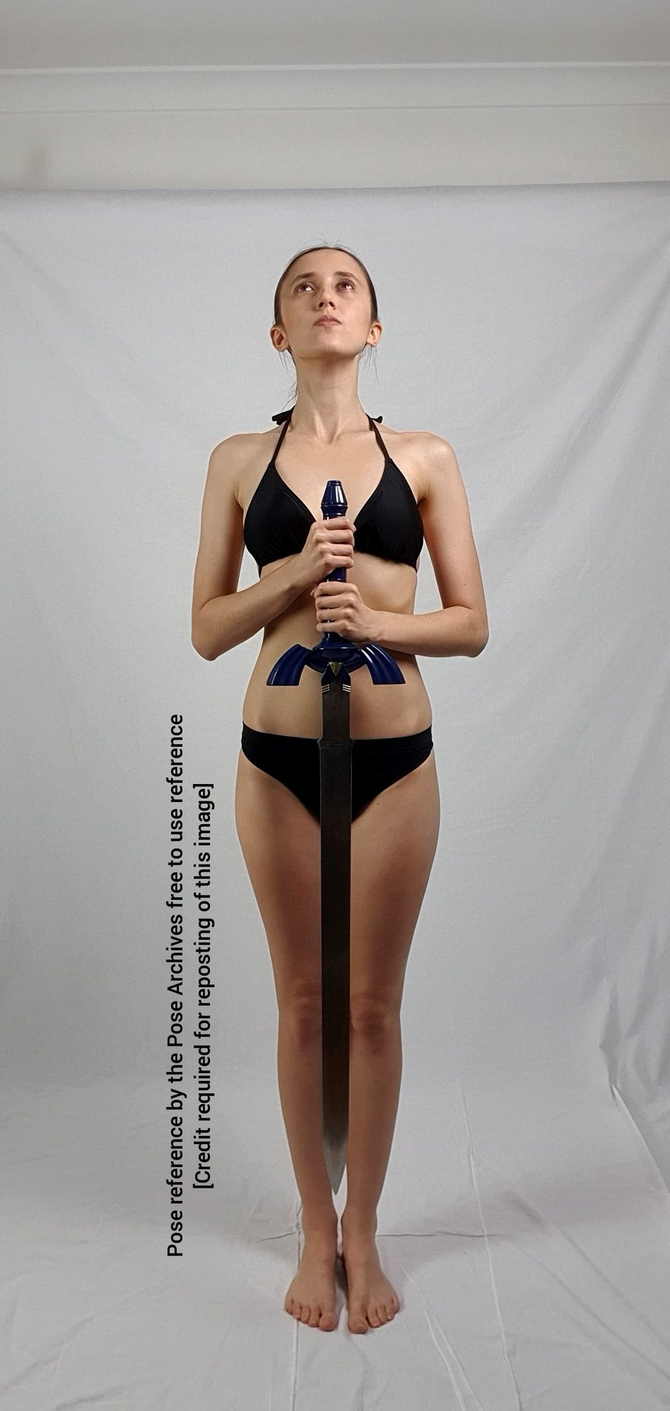 a woman in a black bikini stands with her feet together, holding a large blue and silver sword upright in front of her with both hands. she is looking up, her expression neutral. she is positioned in front of a white backdrop, likely in a studio setting. the overall image is welllit with even lighting.