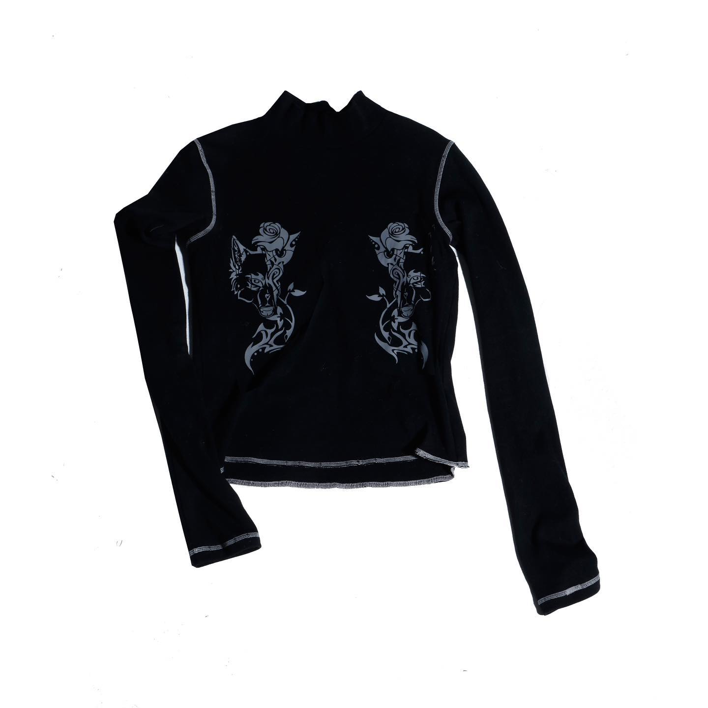 the image shows a black longsleeved shirt with a mock neck. the shirt features a gray print of stylized wolves intertwined with roses, mirroring each other on the front. the fabric appears to be ribbed, and the shirt has a fitted silhouette. the overall style is reminiscent of gothic or alternative fashion, characterized by its dark color and edgy design elements. the shirt is folded and laid flat against a white background for display.