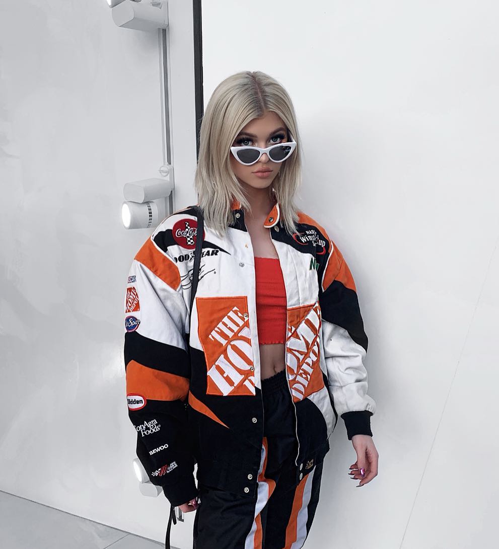 a woman with blonde hair is wearing a black, white, and orange racingstyle jacket with logos and patches, paired with matching black and orange pants. she is also wearing white cateye sunglasses and a red crop top. she is posing against a white wall.
