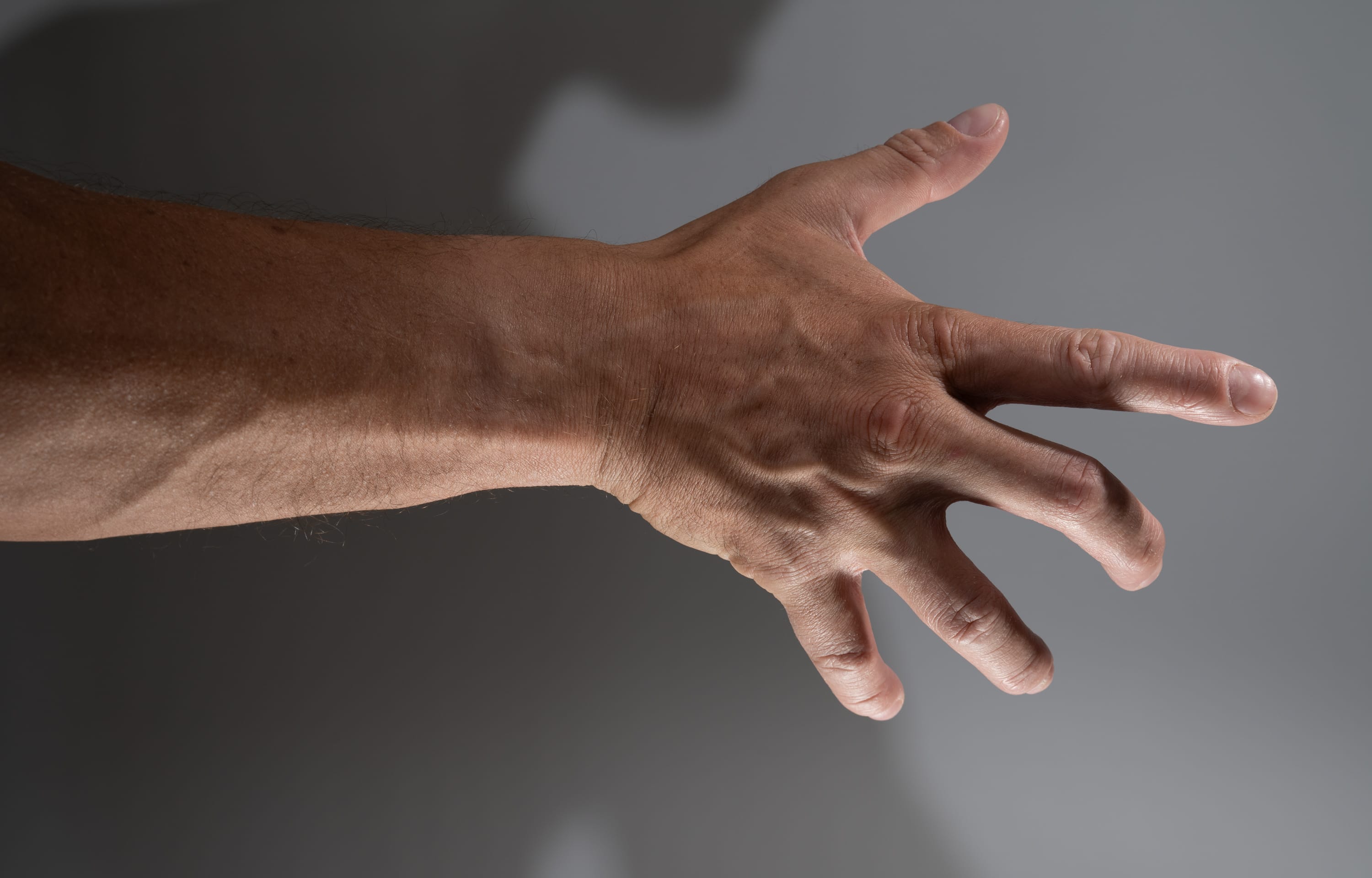a man's arm and open hand are extended against a gray background, casting a shadow. the hand is positioned with the palm facing the viewer, fingers spread. the veins on the arm and hand are visible, highlighting the contours and texture of the skin.