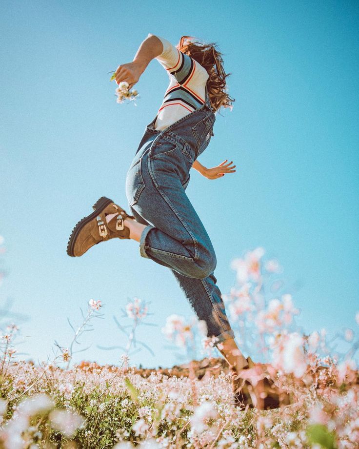 a young woman with long, dark hair is jumping in the air in a field of white flowers. she is wearing a blue denim jumpsuit over a striped shirt, and brown boots. the sky is blue and the sun is shining. the woman is looking forward, and she is smiling. the image is taken from a low angle, so that the woman appears to be very tall. the flowers and the sky are blurred in the background.