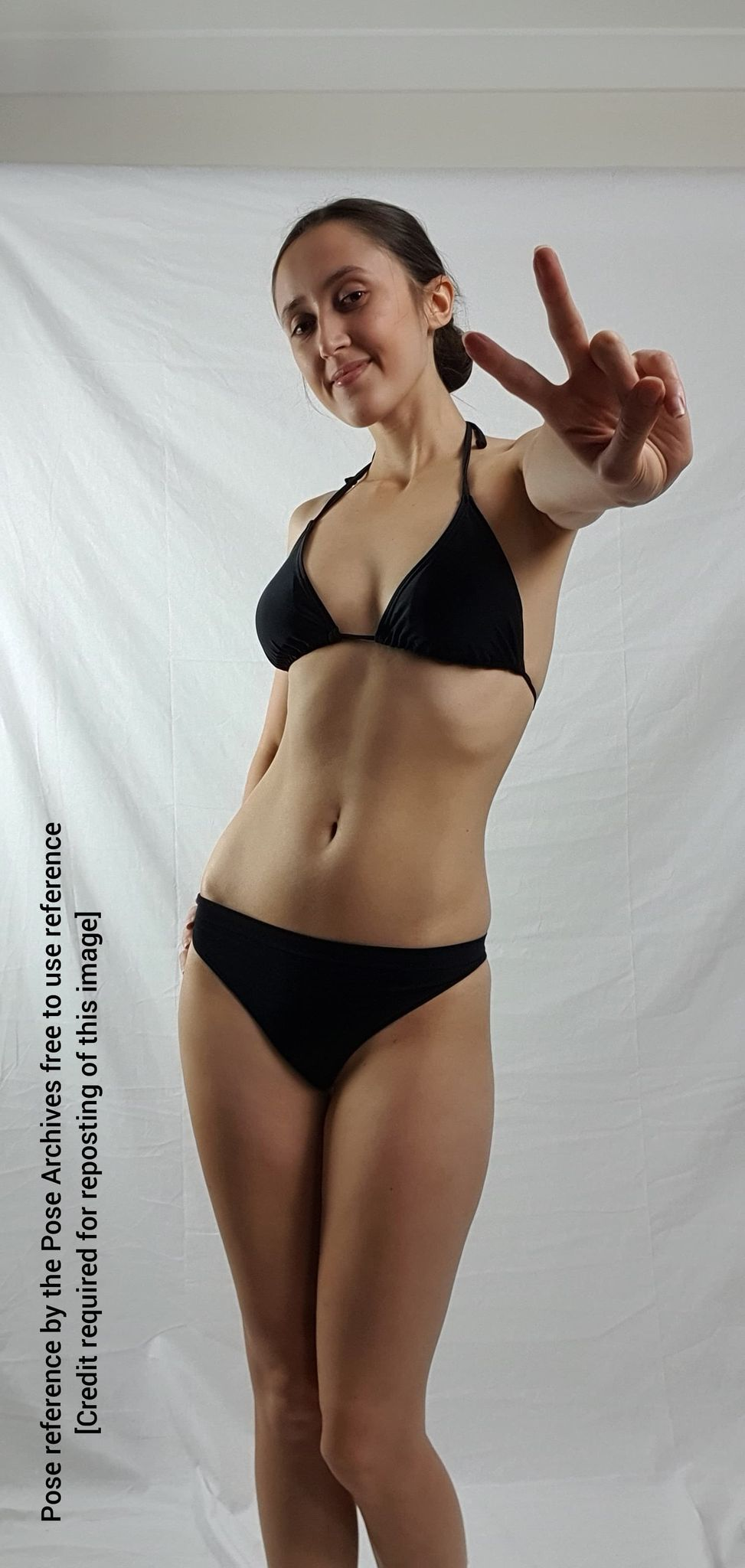 a woman in a black bikini is standing in front of a white backdrop. she is making a peace sign with her right hand. the image is a pose reference, free to use with credit.