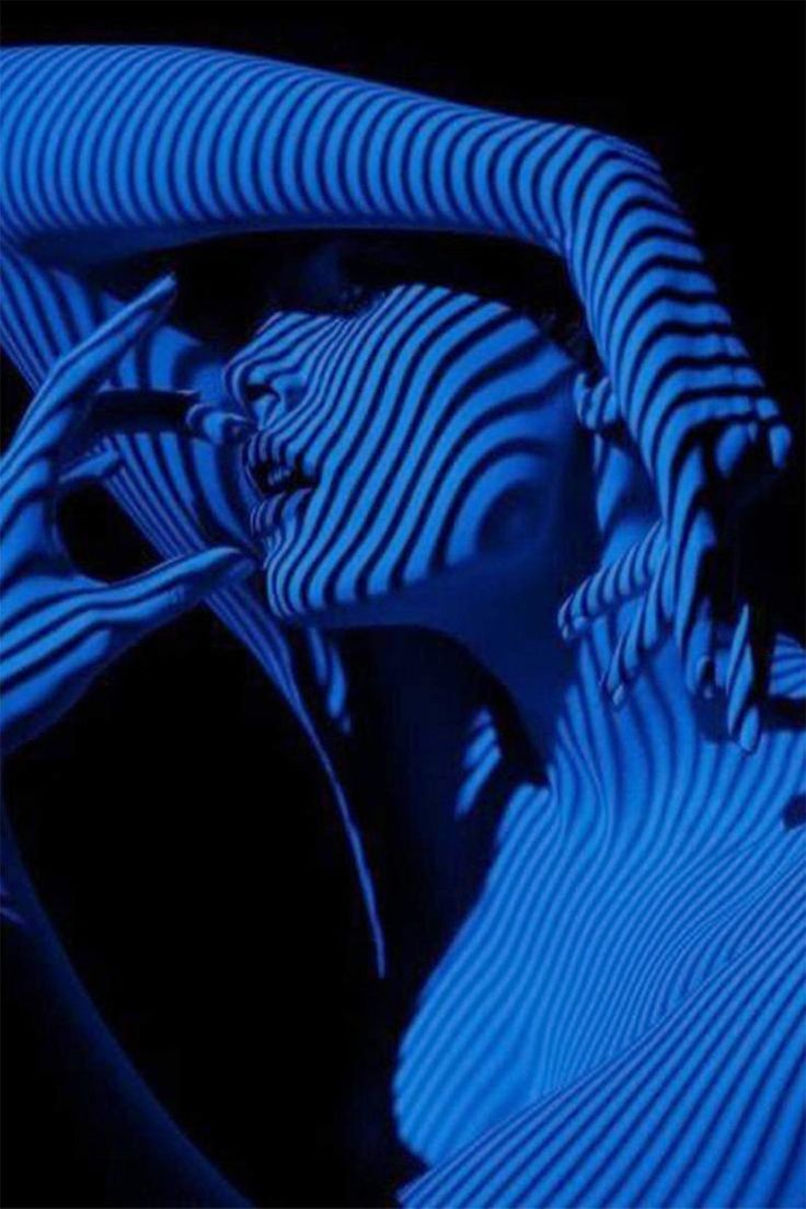 the image is a striking monochrome portrait of a woman, bathed in deep blue light.  her body is dramatically rendered with strong, parallel stripes created by the interplay of light and shadow.  the lines cover her entire body, creating an abstract and almost sculptural effect. the pose is sensual yet mysterious, with hands delicately framing her face. the overall mood is dark, dramatic, and intensely visual, emphasizing texture and contrast. the stark blue enhances the graphic quality of the image and creates a visually arresting piece of art.
