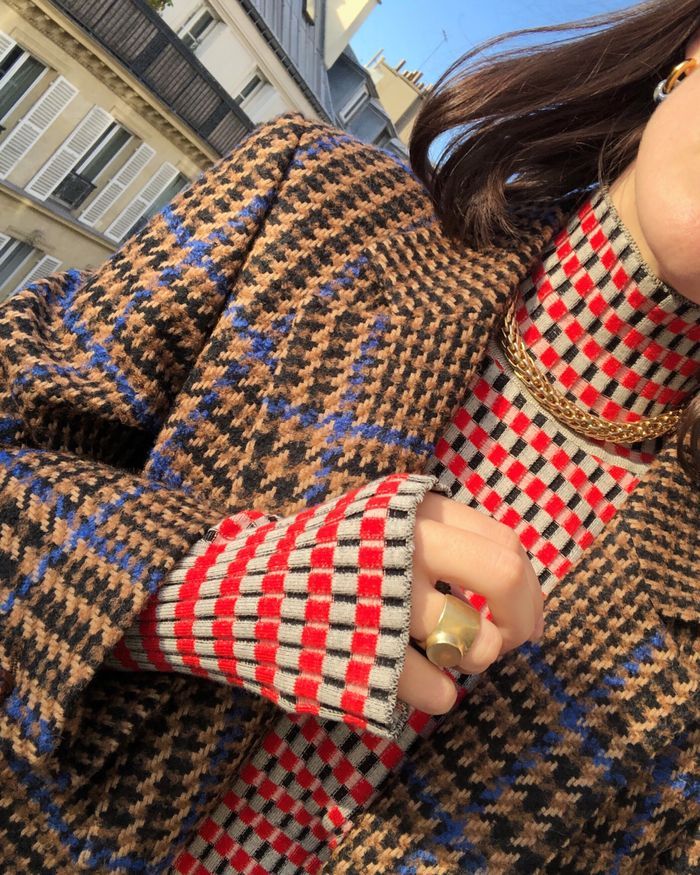 closeup shot of a woman's outfit featuring a brown, blue, and white houndstooth coat. the coat is worn over a red and white checkered turtleneck sweater.  the woman is wearing a gold necklace and a chunky gold ring. her hand is visible, holding the sleeve of the coat. the background is slightly out of focus and shows a parisian street. the overall impression is one of stylish fall or winter fashion.