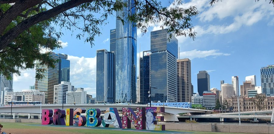 the image shows a cityscape of brisbane, australia, under a partly cloudy sky. the skyline is dominated by several modern skyscrapers reflecting the blue sky. a colorful "brisbane" sign is visible in the foreground, with the victoria bridge and the brisbane river situated behind it. part of a tree frame the upper left side of the image.
