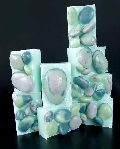 a collection of handmade soap bars shaped like pebbles, stacked in a pyramid shape, featuring a light green and blue color scheme. the soap is designed to mimic the look of natural rocks, creating a visually appealing and spalike presentation. the soap is likely intended for use in bathing or showering, offering a luxurious and aromatic experience.