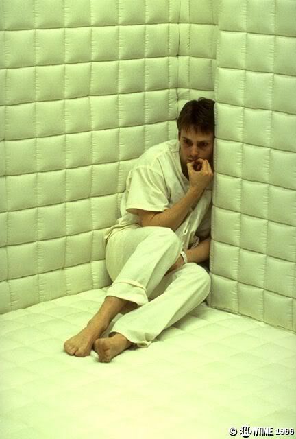a man in a white padded room, dressed in white, sits in a corner of the room, looking anxious and distressed. the walls are padded with white squares, creating a sterile and unsettling atmosphere. the man's posture and expression suggest he is in a state of distress, potentially in a mental health institution or undergoing therapy. the image evokes a sense of isolation and fear, characteristic of scenes depicting mental health struggles.