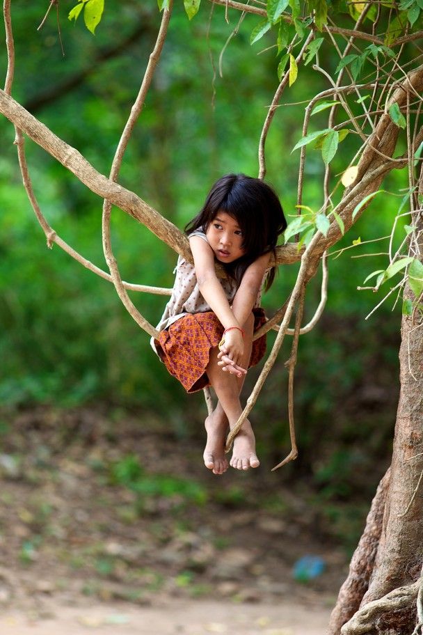 a young asian girl sits barefoot in the crook of a tree branch, her legs dangling. she wears a simple top and a patterned skirt. her dark hair frames her face as she gazes off to the side with a pensive expression. the scene is set outdoors, surrounded by lush green foliage and branches. the overall mood is one of serenity tinged with a hint of sadness.