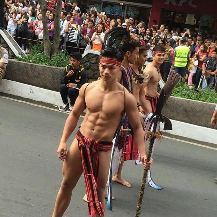 the image shows a group of shirtless, muscular filipino men participating in a parade or festival. they are wearing traditional tribal clothing, including bahag (loincloths) and carrying spears.  the men are the focus of the image, with a large crowd of spectators visible in the background. the setting appears to be a city street during a cultural celebration or event. the overall atmosphere suggests a vibrant and proud display of filipino heritage and indigenous culture.