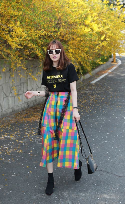 a young woman is walking down a street lined with yellow forsythia bushes. she is wearing a black tshirt with "nebraska athletic dept" on it, a colorful checkered midi skirt with a black lace overlay and zipper detail, white sunglasses, and black ankle boots. she is carrying a small black handbag. the overall style is casual and springlike. the woman appears to be of asian descent.