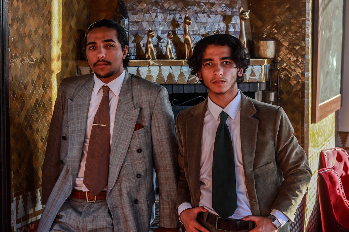 two stylishly dressed men are captured in a full shot, seated and posing confidently.  they are wearing classic suits; one in a gray plaid suit and the other in a brown corduroy suit. both men have a polished and puttogether look. the setting is an interior room with a distinctive moroccanstyle tiled wall and fireplace, adding a vintage or retro aesthetic.  the overall mood is elegant and sophisticated.