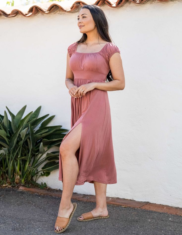 a woman in a pink dress is standing outdoors, smiling. she is wearing sandals and her hair is down. there is a wall behind her and some plants to the left. the lighting is natural and sunny.