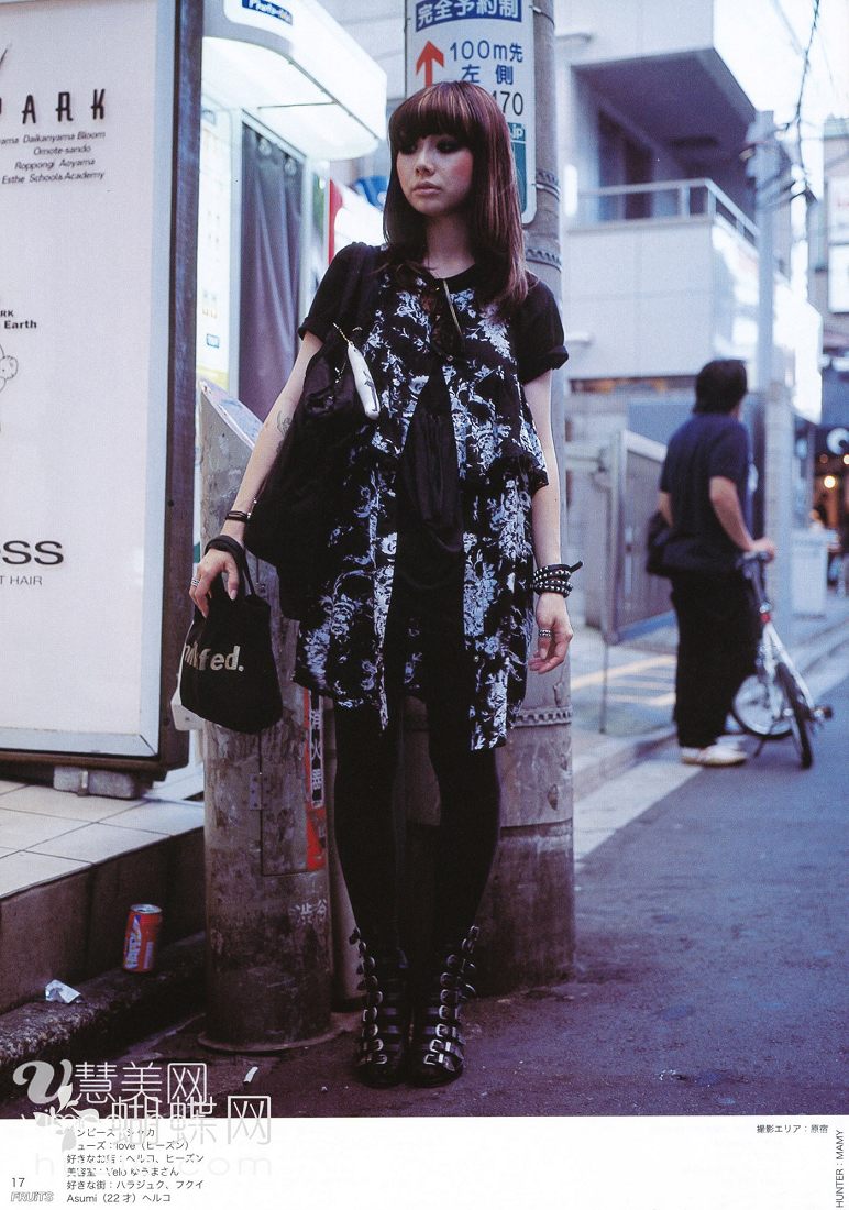 a young woman with dark hair and dark clothing stands on a city sidewalk, posing for the camera. she wears a black and white patterned dress, black leggings, and black buckled boots. she carries a black bag and has a white shopping bag with "fed." printed on it hanging from her wrist. the background features a man walking away from the camera and a street scene with buildings and signs written in japanese. the overall color palette is dark and muted, with a slightly overcast daylight illuminating the scene.