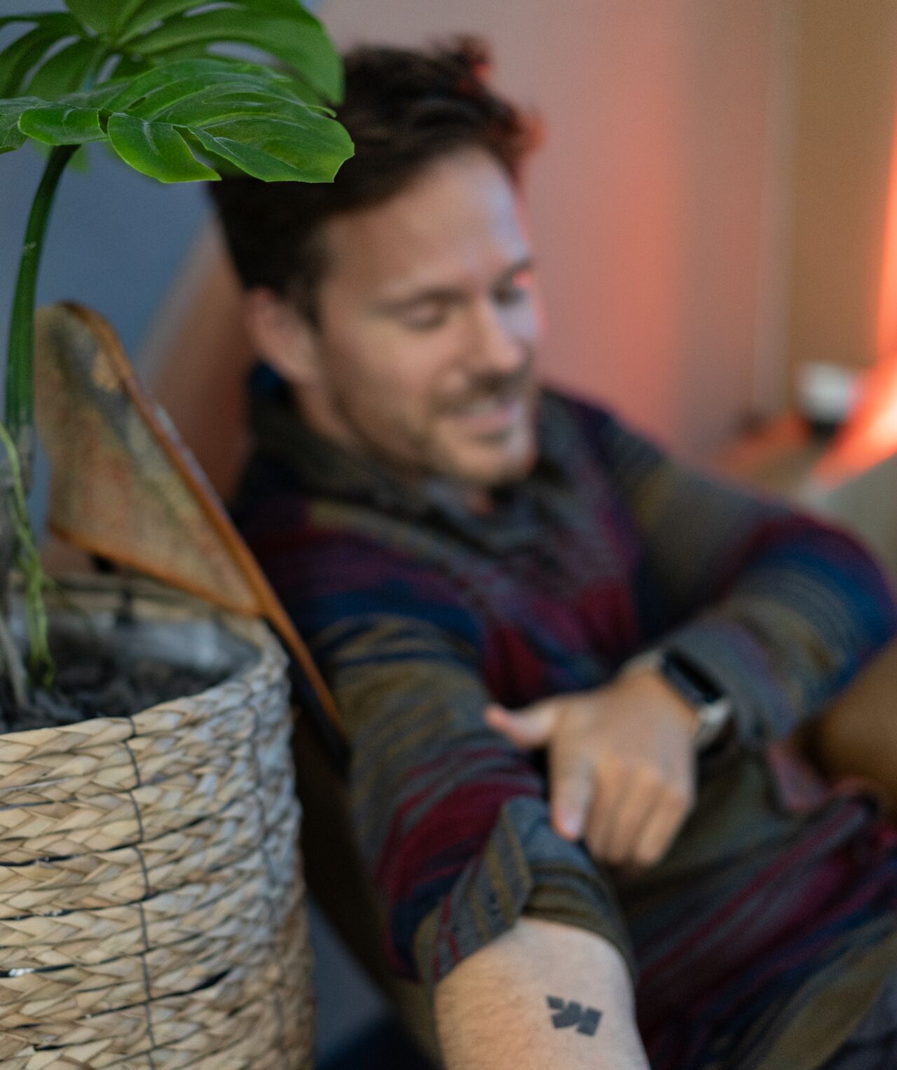 the image shows a closeup of a man's arm, which features a small, simple tattoo. he's sitting casually, wearing a multicolored sweater, in what appears to be a relaxed and comfortable home environment.  the background is slightly out of focus, showing a warm, orangetoned room.  a large monstera deliciosa plant in a wicker basket is prominently featured in the foreground to the left, partially obscuring the man's torso.  the overall mood is peaceful and intimate.