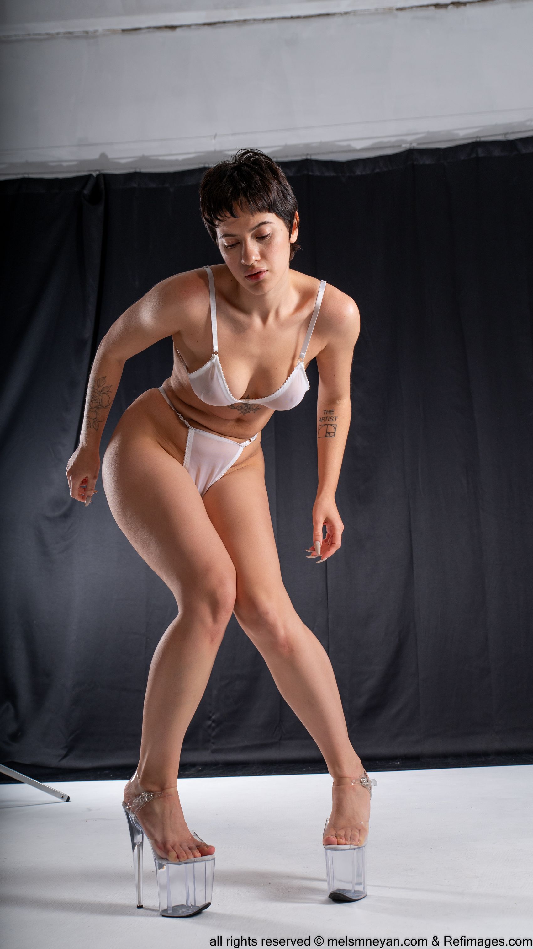 a woman stands in a studio wearing white lingerie and clear heels. she has short hair and tattoos on her arms. she is looking down.