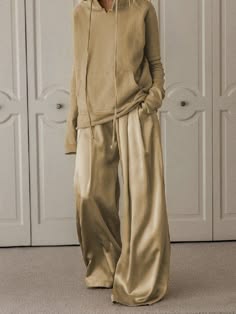 a woman in a beige outfit stands with her back to a white wall. she is wearing widelegged pants and a longsleeved sweater with a hood. she is standing in a room with white walls and doors.
