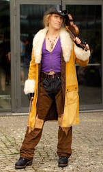 a full shot of a male cosplayer dressed as a cowboy character. he is wearing a long yellow coat with a faux fur collar, brown chaps, a purple shirt, and a cowboy hat. he is holding a rifle and is standing on a street in front of a building.