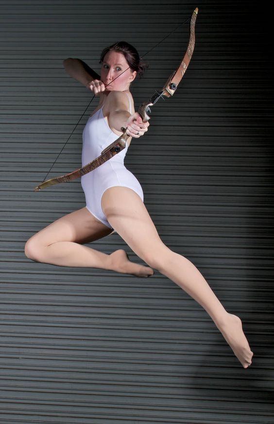 a woman in a white leotard is captured midair, executing a powerful jump while aiming a bow and arrow. the studio setting provides a neutral backdrop, allowing the focus to remain on the woman's athleticism and the dynamic pose. the image emphasizes the strength and grace required for archery.