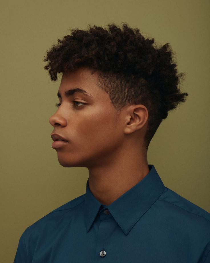 a young man with curly black hair and a blue shirt looks to the left side of the frame in a profile shot. he is standing in front of a muted green background with soft light. he has a cleanshaven face with a subtle smile playing on his lips.