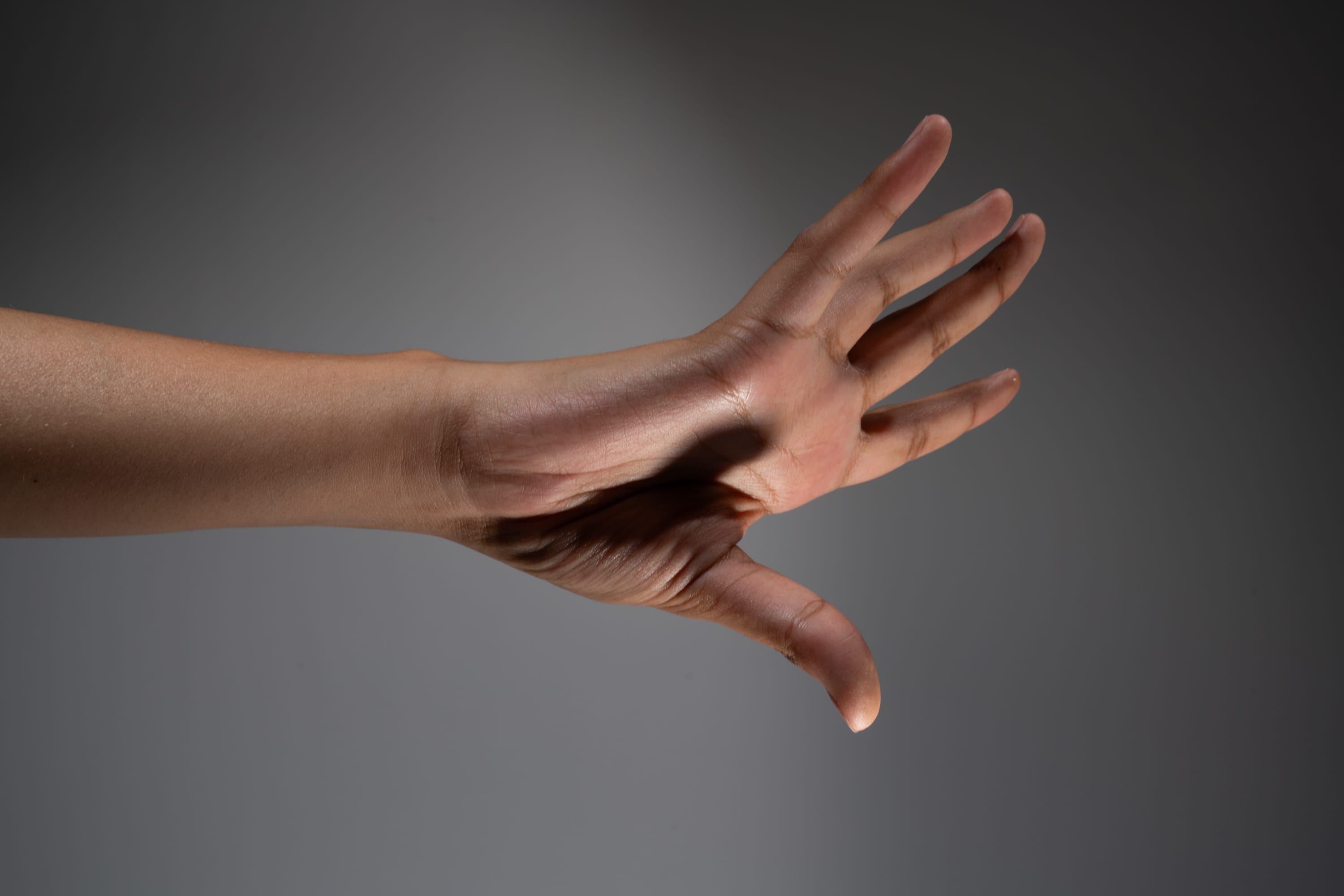 a closeup image of a person's hand and arm extended against a gray background. the hand is open, with the fingers spread slightly apart. the image is lit with studio lighting, creating a soft shadow on the arm.