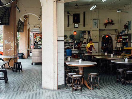 a view of a cafe's outdoor seating area located in a city. there are several tables and chairs set up outside, with customers enjoying food and drinks. the cafe has a rustic and traditional look, with wooden tables and chairs, and a tiled floor. the cafe is located in a building with a white facade and a covered entrance, creating a sheltered seating area. the cafe is open to the street, allowing customers to enjoy the city atmosphere. the image captures the cafe's ambience and the hustle and bustle of city life.