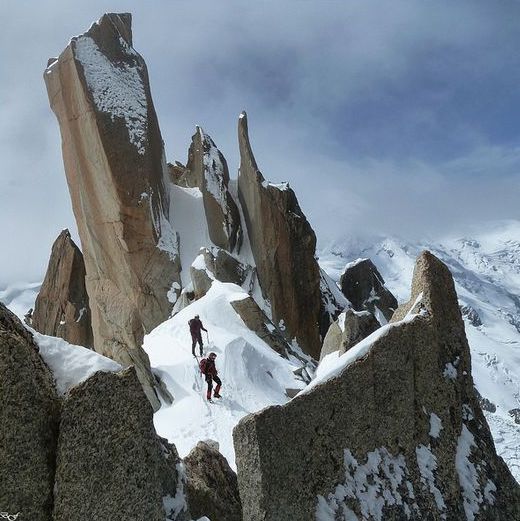 a scenic image shows two climbers navigating a snowy, rocky mountain terrain. the landscape is dominated by sharp, jagged rock formations covered in snow, with a larger mountain range visible in the background under a cloudy sky.