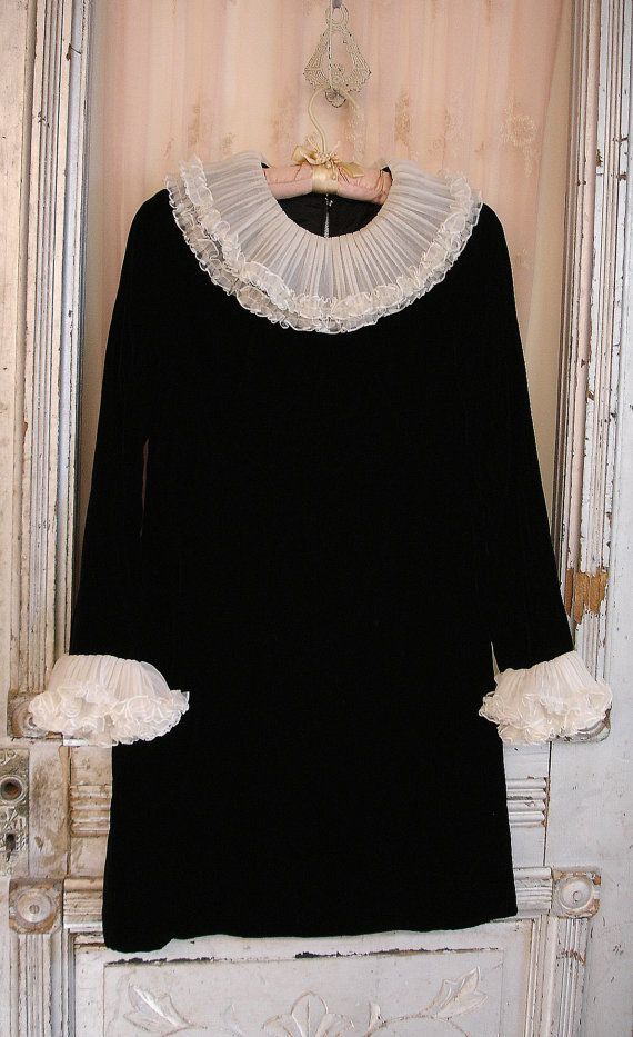 a black velvet dress with a white lace collar and cuffs, hanging on a hanger. the dress is a vintage style with a simple silhouette. it's hanging against a background of a white door, with a vintage white door knob.