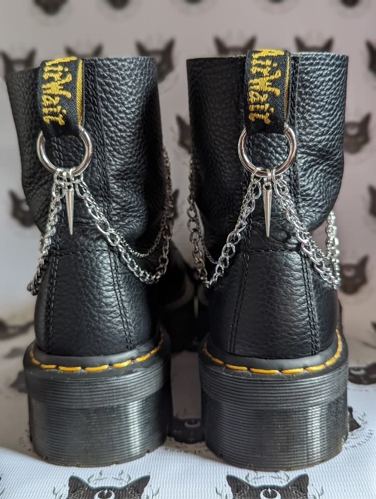 a pair of black leather platform boots with chains attached to the ankles. the boots are a grunge/punk style, with a chunky platform sole and a black leather upper. the chains are silver and are attached to the ankles of the boots, with a couple of buckles. the boots are edgy and cool, and would be a great statement piece for any grunge/punk outfit.