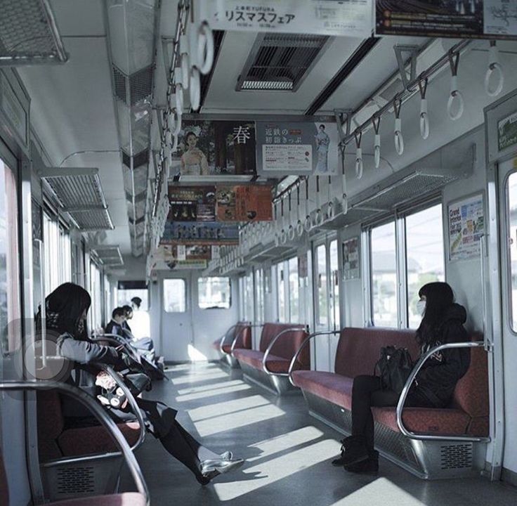 the image shows the interior of a japanese train car.  sunlight streams in through the windows, illuminating the red seats and the few passengers. two women sit in the foreground, one engrossed in something in her lap and the other looking out the window. other passengers are seated further down the car.  the overall atmosphere is quiet and peaceful.  numerous advertisements are visible on the walls of the train.