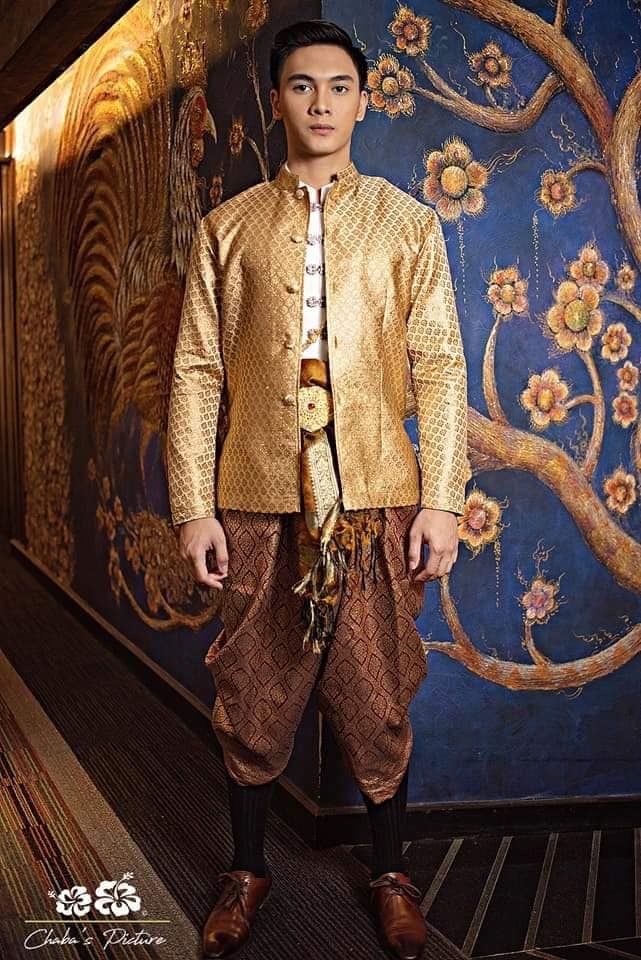 a man in a traditional thai costume is standing in front of a wall with floral art.  he is wearing a gold jacket with a brown sash and brown patterned pants.  he is also wearing black socks and black shoes. he is standing in a formal pose and looking directly at the camera.  the wall behind him has a blue background with gold and brown floral patterns.  this image is a portrait of a man in traditional thai clothing.