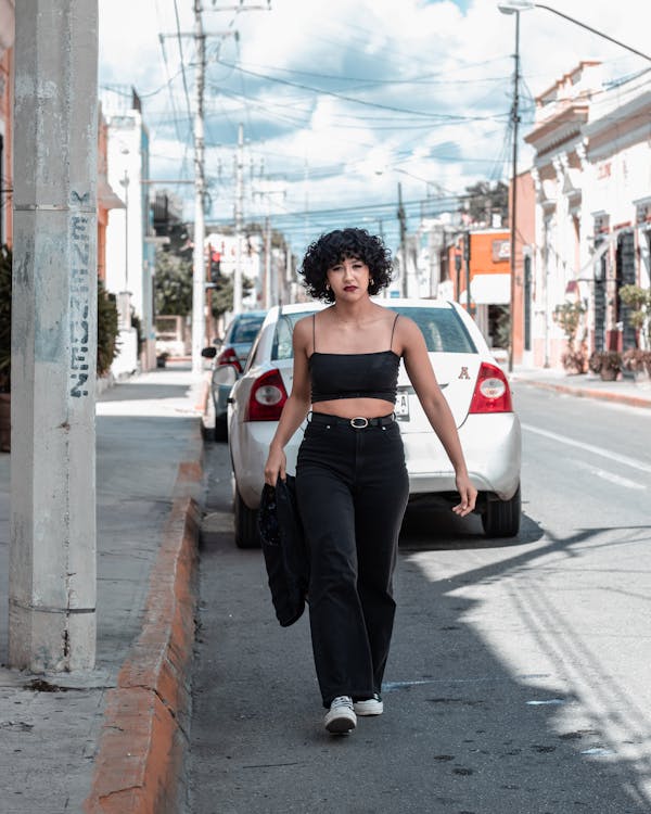 a young woman with curly dark hair walks down a city street, carrying a black bag. she is wearing black pants, a black crop top, and sunglasses.  parked cars are visible along the street, and buildings line the road. the overall scene is casual and urban, bathed in sunlight. the focus is on the woman and her confident demeanor.