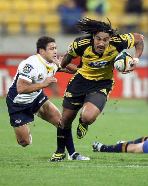 a rugby player in a yellow and black jersey is running with the ball in his hands while being tackled by a player in a white and blue jersey. the player with the ball is wearing black shorts and yellow and black socks while the other player is wearing blue shorts and yellow socks. the game is being played on a green field.