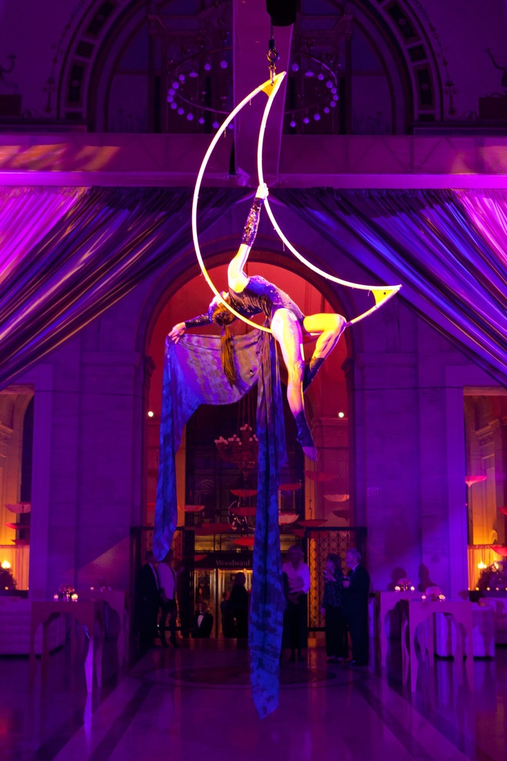 a woman in a black leotard is performing an aerial dance on a crescent moonshaped hoop suspended from the ceiling of a large venue. she is holding onto the hoop with both hands and her feet are dangling. the hoop is lit up with a golden light and the woman's silhouette is clearly visible against the purple lighting. the background is blurry and out of focus but there appears to be people watching the performance. the venue has tall ceilings and large windows. the woman is graceful and elegant in her movements and the performance looks impressive.