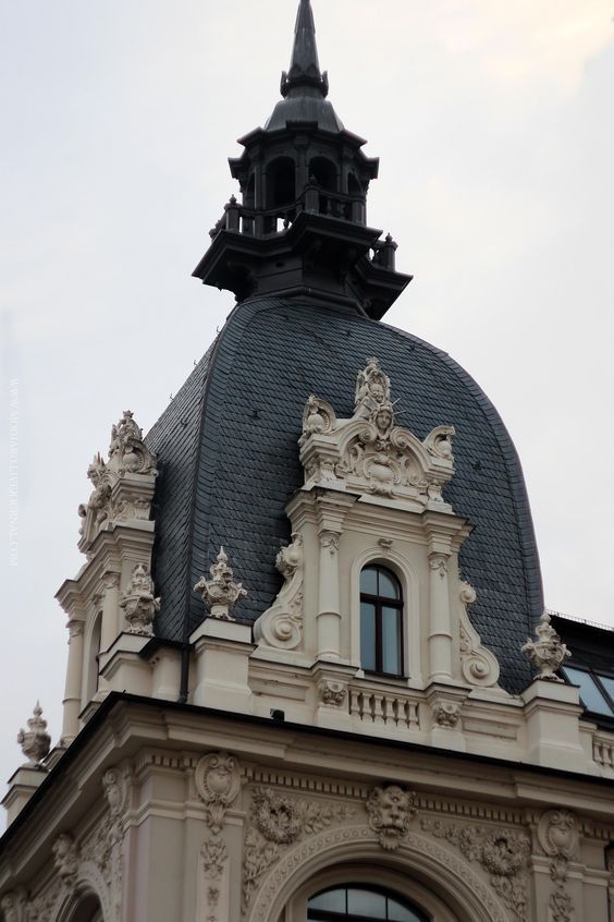 loseup view of the ornate architectural details of a building's rooftop. the image shows a dark grey slate dome with a tall, black spire, surrounded by intricately carved stonework.  the building is predominantly light beige or offwhite, with numerous sculptures and relief carvings embellishing the facade. a window with a arched top is visible under the dome. the style appears to be classical or european, possibly from the late 19th or early 20th century. the sky is overcast and a muted white.