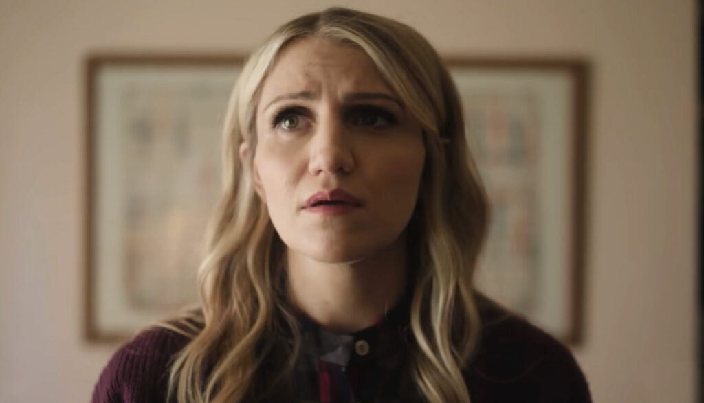a closeup portrait of a woman with blonde, wavy hair looking upwards with a concerned expression. she is wearing a darkcolored collared shirt with a button and a darkcolored sweater. in the background, there is a framed artwork hanging on the wall. the lighting is soft, casting shadows on her face.
