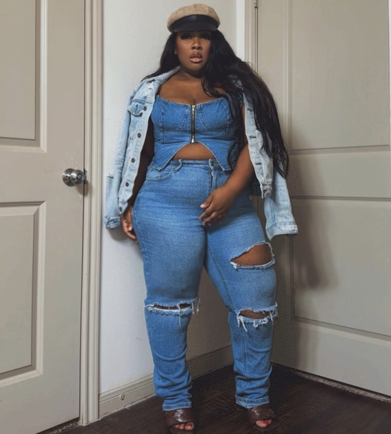 a woman wearing a blue denim crop top corset, ripped jeans, and a jean jacket. she is also wearing a light brown newsboy cap and brown heeled sandals. she is standing in a doorway with her hands on her hips and her legs crossed.