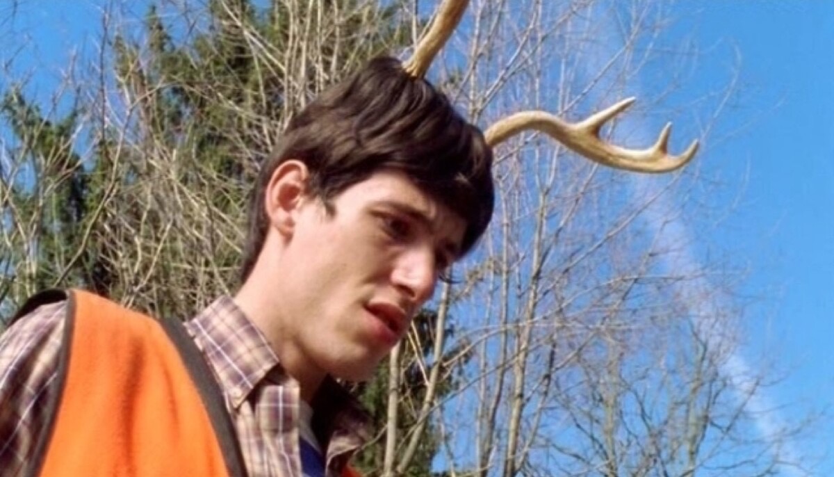 a man with antlers on his head is smiling happily while standing in a forest. he is wearing a plaid shirt and an orange vest. the trees are bare and the sky is blue. it is a sunny day. he is enjoying a day out in nature.