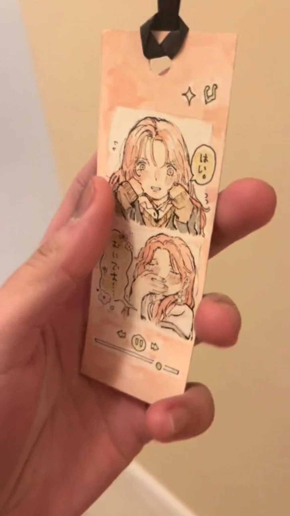 a hand is holding a pink bookmark with an anime drawing of two young girls in a cartoon style.  the bookmark is made of paper with a brown strap. one of the girls is wearing a pink coat while the other is in a green shirt with a light brown jacket. the drawing is in a pink cartoon style.