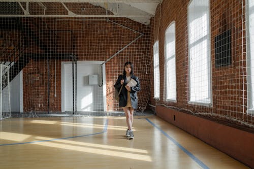 a young asian woman stands alone in a large gymnasium with brick walls and large windows. she is wearing a black jacket and a beige skirt and holding a book. sunlight streams through the windows, casting shadows on the floor.  a net is visible along the wall. the overall atmosphere is quiet and contemplative.