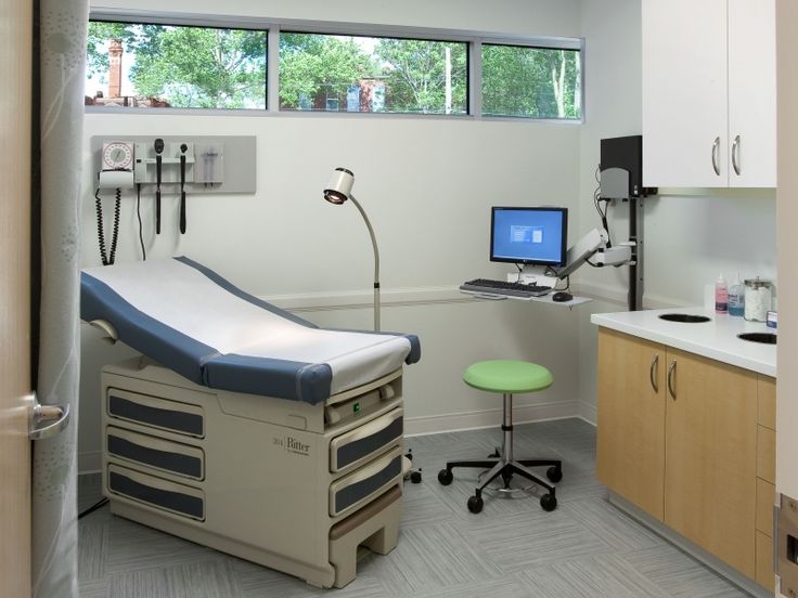a modern medical examination room with an exam table and a chair, cabinets with drawers, a small desk and computer, and a lamp.