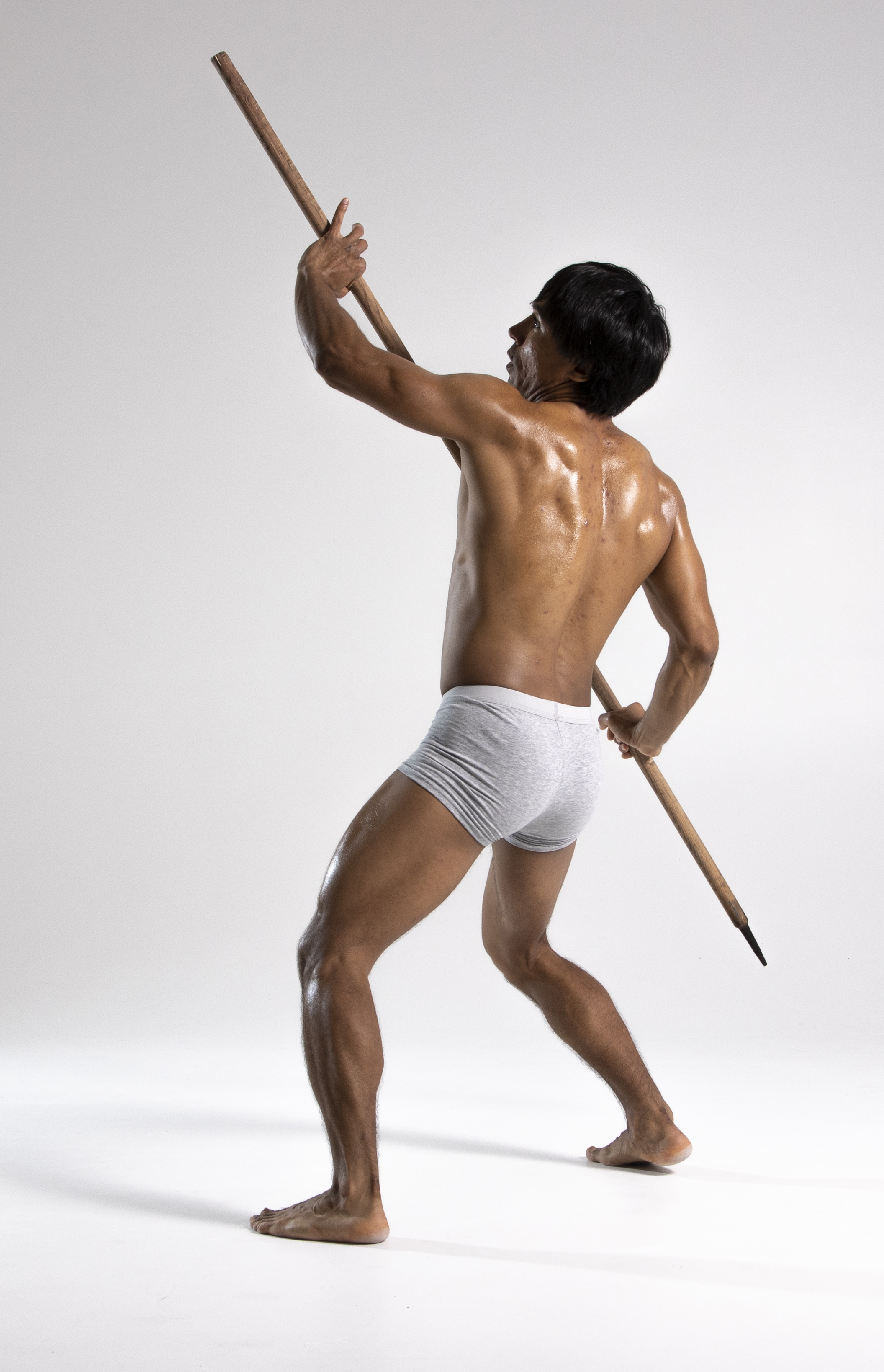 an athletic, barechested man wearing gray underwear stands with his back to the viewer, holding a long pole or spear upwards. he has muscular arms and legs and his back is slightly sweaty. he is looking up toward the top of the pole. the background is a plain white studio setting.