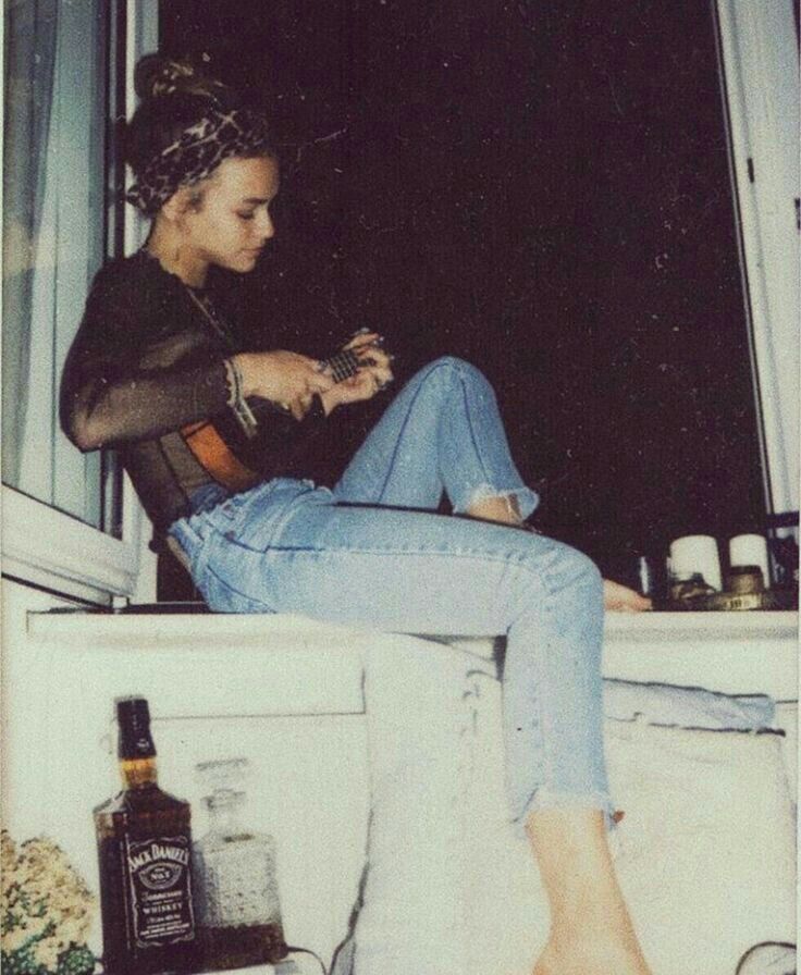 a young woman in a black shirt and blue jeans sits by a window playing a guitar at night. there is a bottle of jack daniels on a table beside her. the photo has a vintage, retro feel.