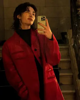 a young man with long dark hair takes a selfie in a red leather coat. he's standing on a staircase inside a building, and the photo has a moody, stylish aesthetic.  the red coat is the focal point of the image, and the man's expression is serious and somewhat mysterious. the lighting is dim, creating shadows and highlighting the texture of the leather.
