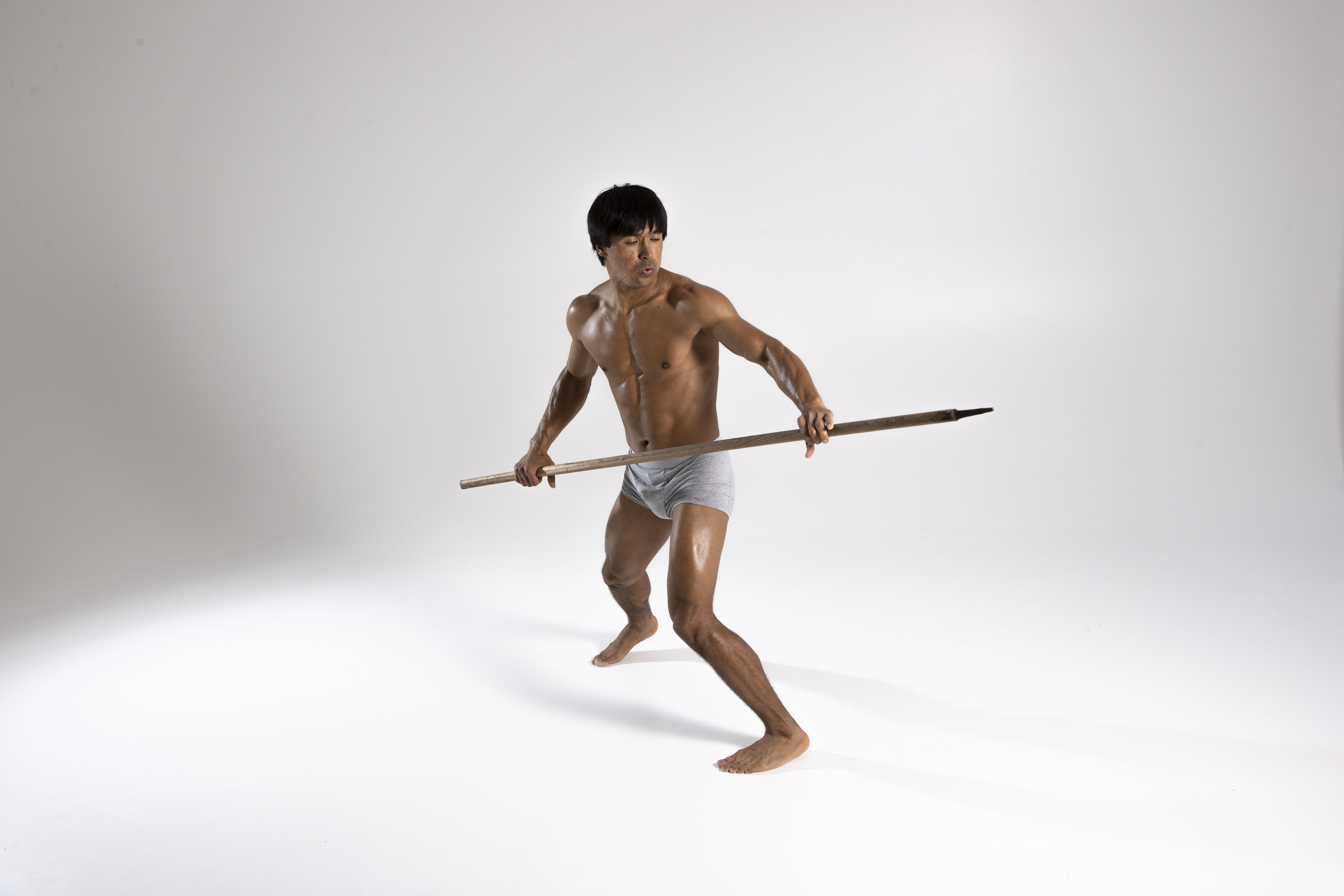 a shirtless asian man wearing underwear stands barefoot in a studio, holding a spear with both hands. he is in a fighting pose, looking off to the side. he has an athletic build. the background is plain white.