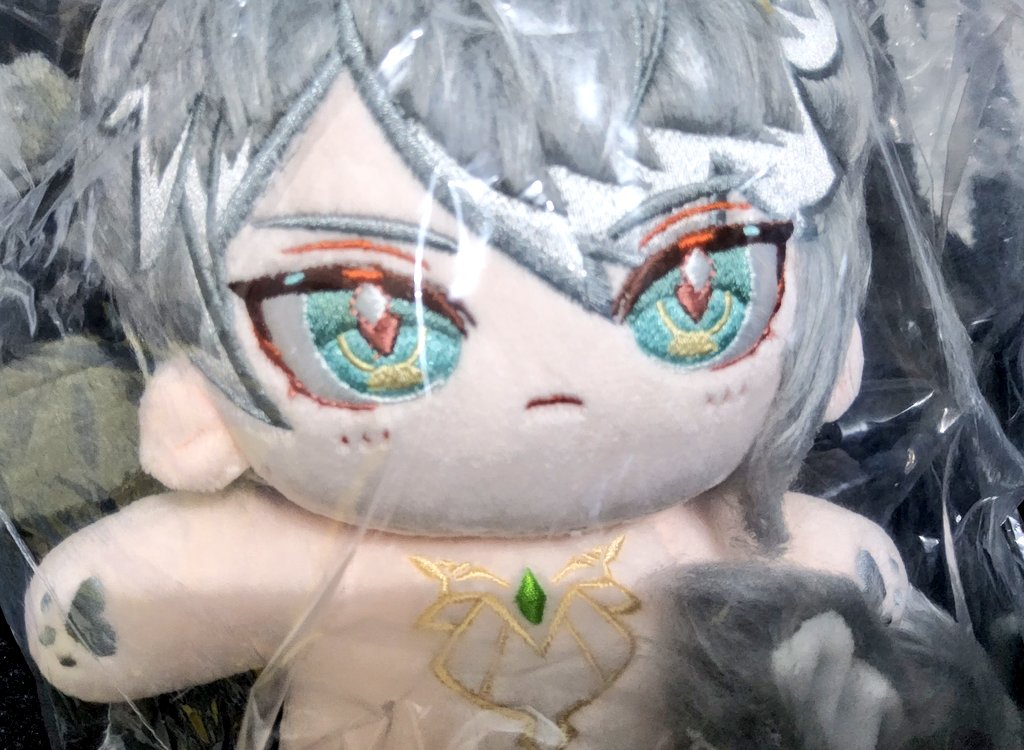 a closeup of an anime plush toy. the plush has grey hair, pink skin, and big blue eyes with gold and green accents.  it's wearing a golden necklace with a green gem. the plush is sitting in plastic wrapping.