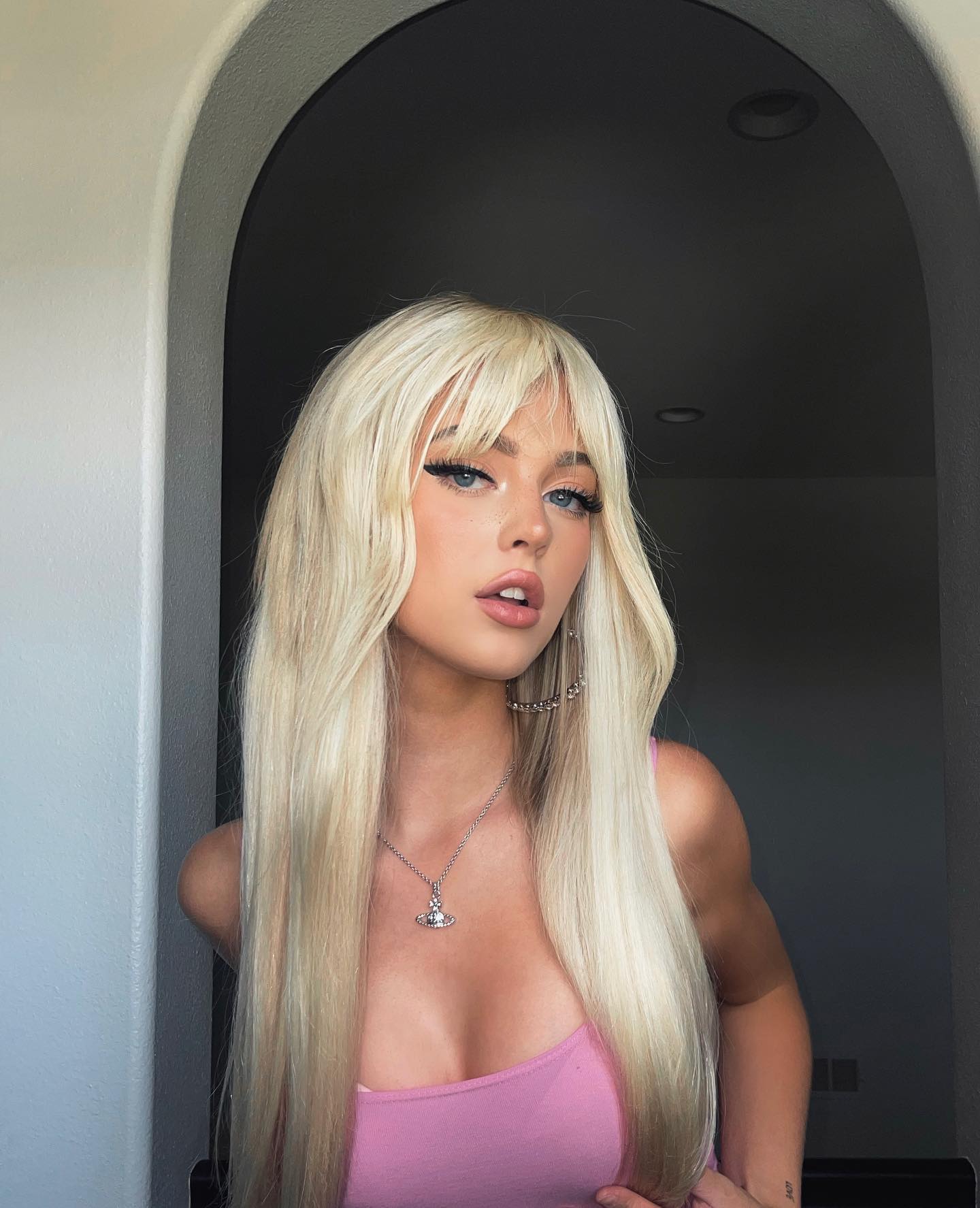 a portrait of loren gray standing in a doorway. she has long, blonde hair with bangs, and blue eyes with dramatic eyeliner. she is wearing a pink tank top and a necklace with a pendant. she also has earrings. she is standing in an archway or doorway with a dark room behind her.