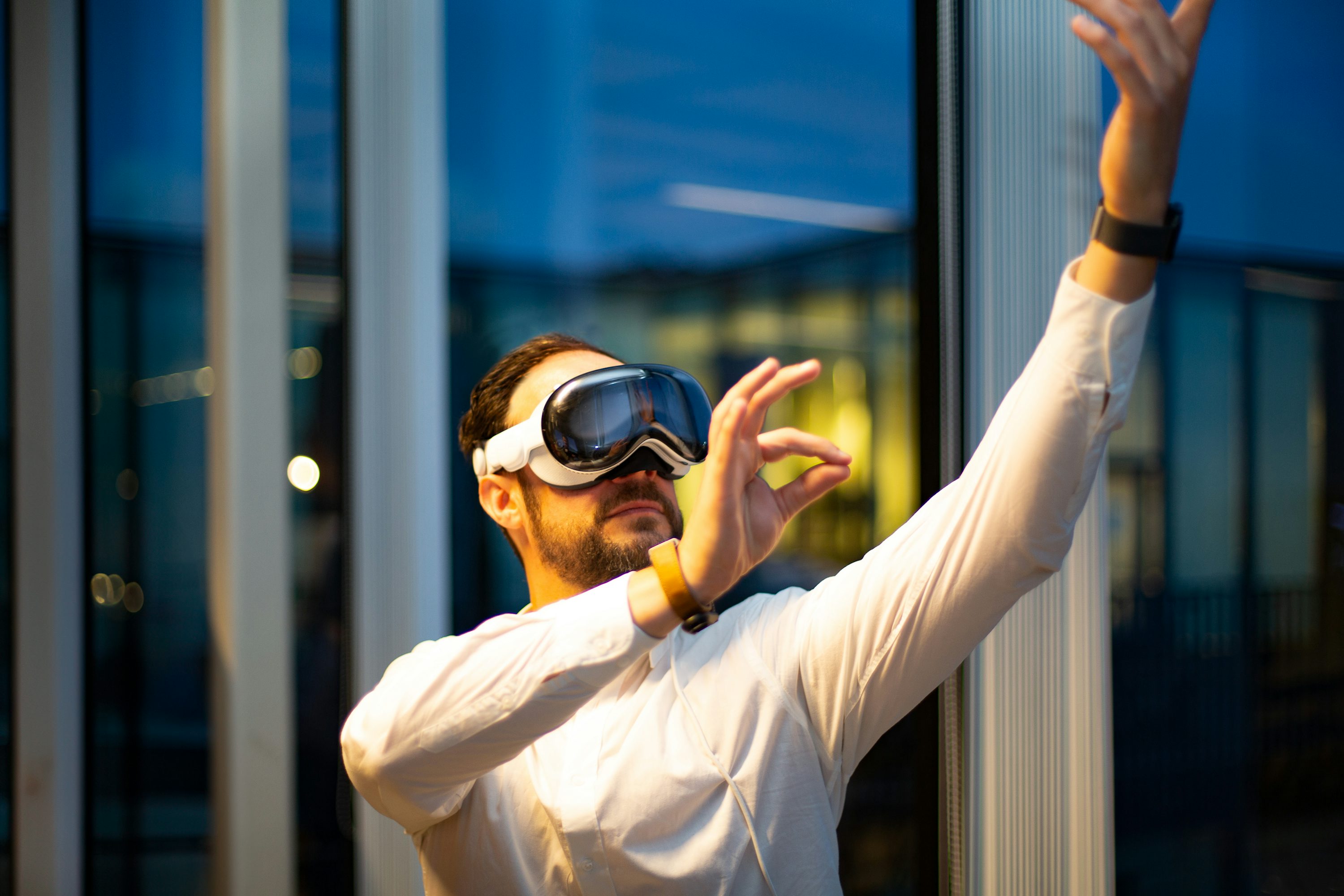 a man wearing a vr headset stands indoors with his arms raised, making gestures. he is wearing a white shirt and appears to be interacting with a virtual environment. the background features a modern interior with glass windows.
